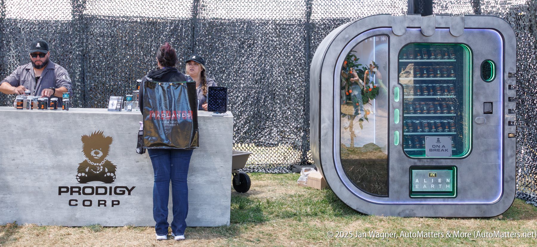 A woman with a large backpack stands at a Prodigy Corp booth next to a futuristic vending machine labeled "Alien Earth" at an outdoor event.
