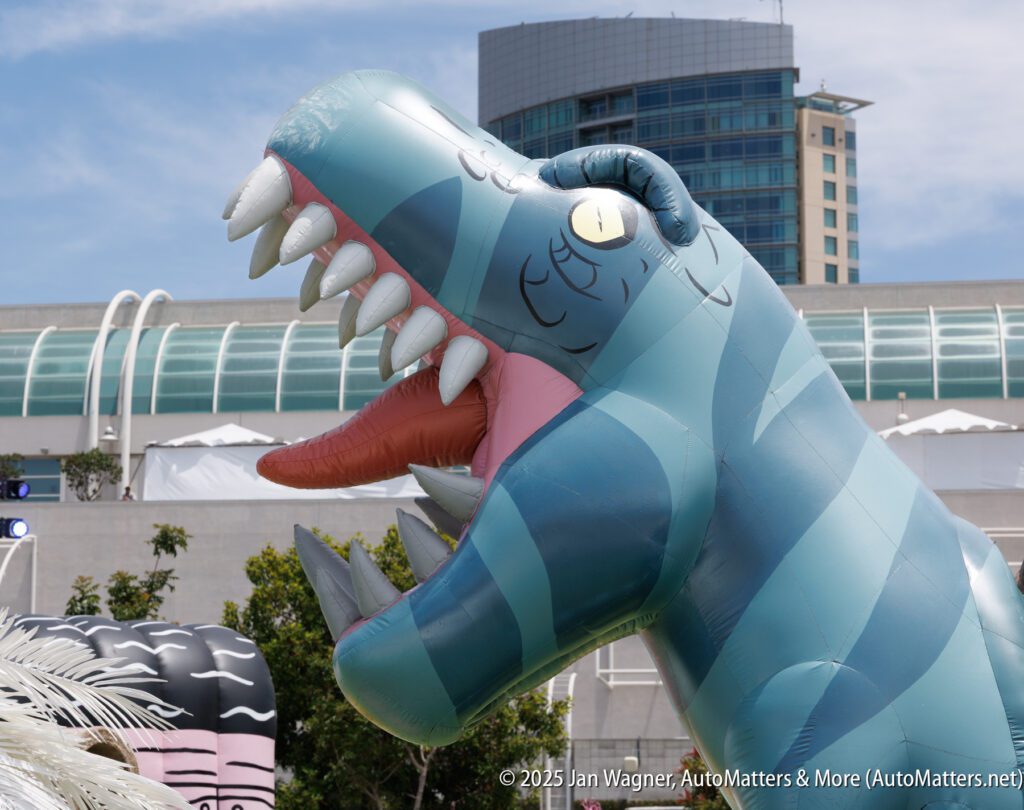 A large blue inflatable dinosaur with sharp teeth and yellow eyes stands outdoors near modern buildings under a blue sky.