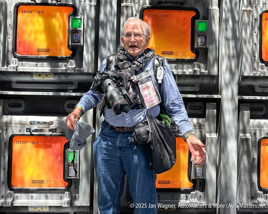 An older man with cameras and press badges stands in front of industrial-looking panels with orange-lit displays.
