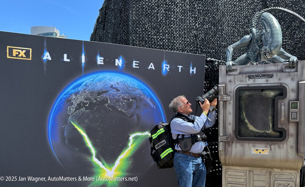 A man takes a photo of an "ALIEN: EARTH" display featuring a large alien figure emerging from a prop structure at an FX promotional event.