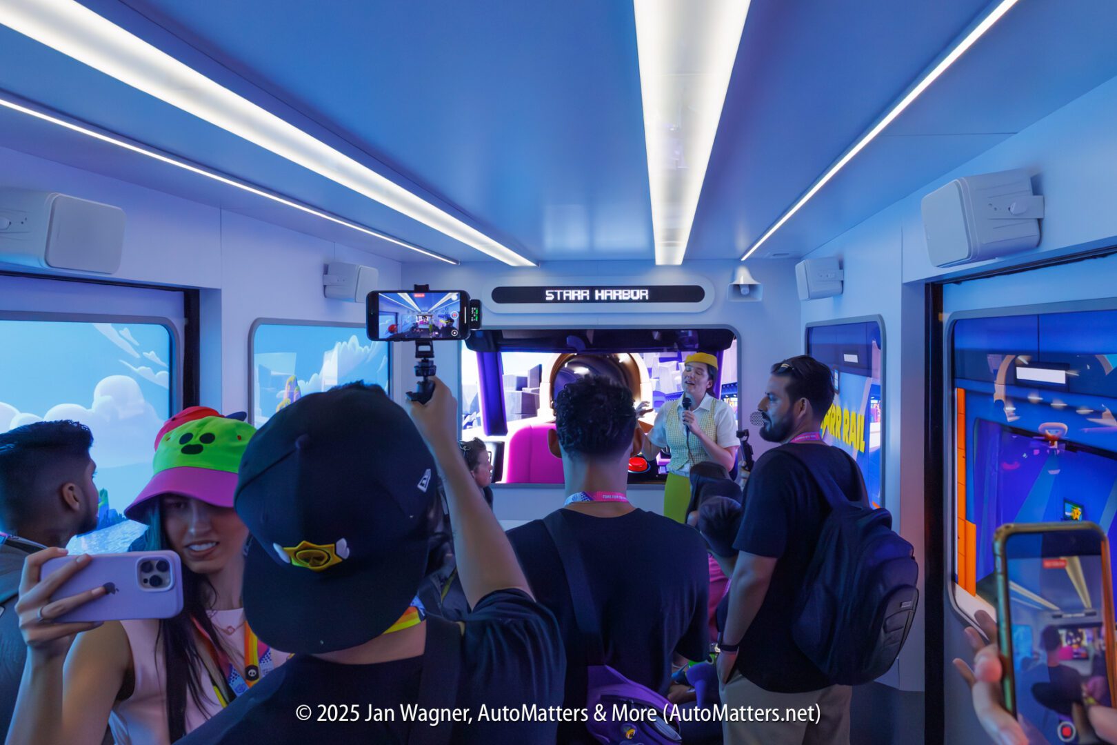 A group of people take photos and videos inside a themed train car labeled "Starhill Harbour," with screens showing colorful animated scenes.