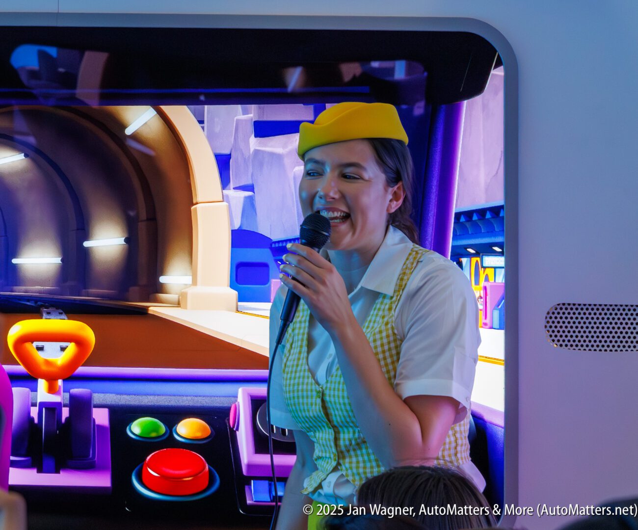 A woman in uniform and yellow hat speaks into a microphone in front of a colorful interactive display with large red and green buttons.