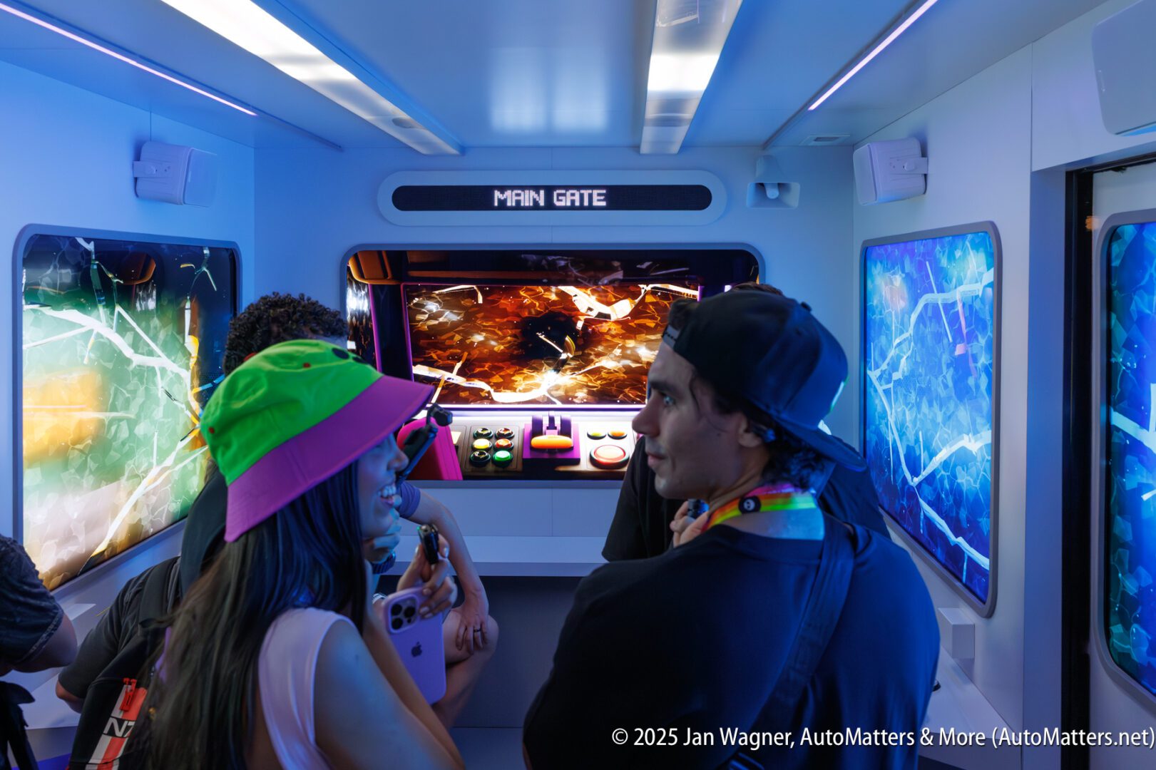 People stand inside a futuristic-themed room with colorful displays and a screen labeled "MAIN GATE," interacting with various consoles and buttons.