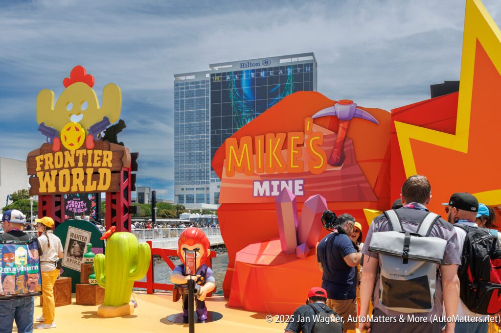 Visitors gather around colorful "Frontier World" and "Mike's Mine" displays at an outdoor event, with a large Hilton hotel building visible in the background.