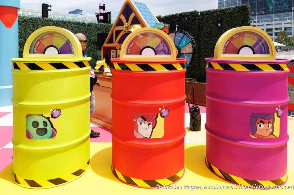 Three colorful barrels, each labeled with a different character face and meter on top, are displayed outdoors at an event with props and booths in the background.