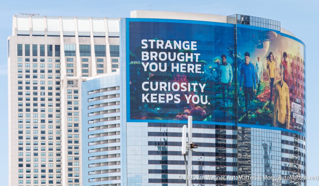 A large outdoor billboard on a modern building displays the text "Strange brought you here. Curiosity keeps you." with images of people and futuristic scenes.