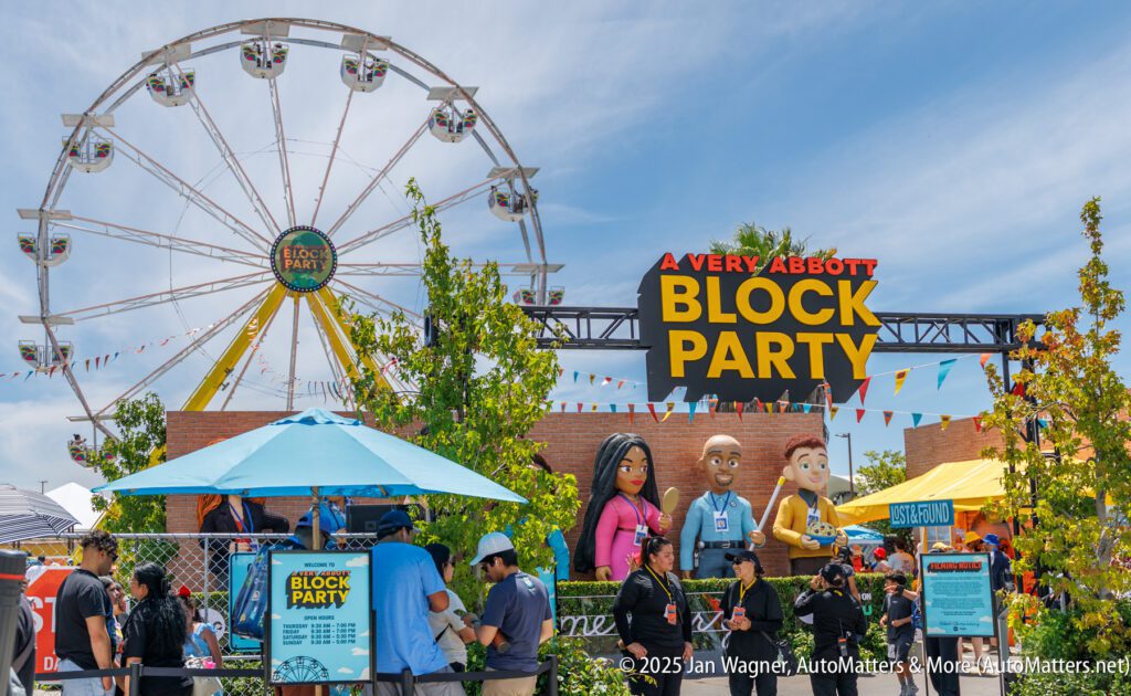 People gather at "A Very Abbott Block Party" event, featuring large character figures, a Ferris wheel, and outdoor decorations under a sunny sky.
