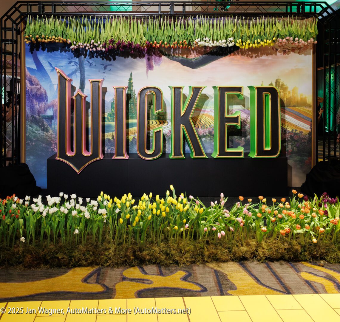 Large "WICKED" sign displayed in front of a tulip garden, with a backdrop of a fantasy landscape and yellow brick road motif on the floor.