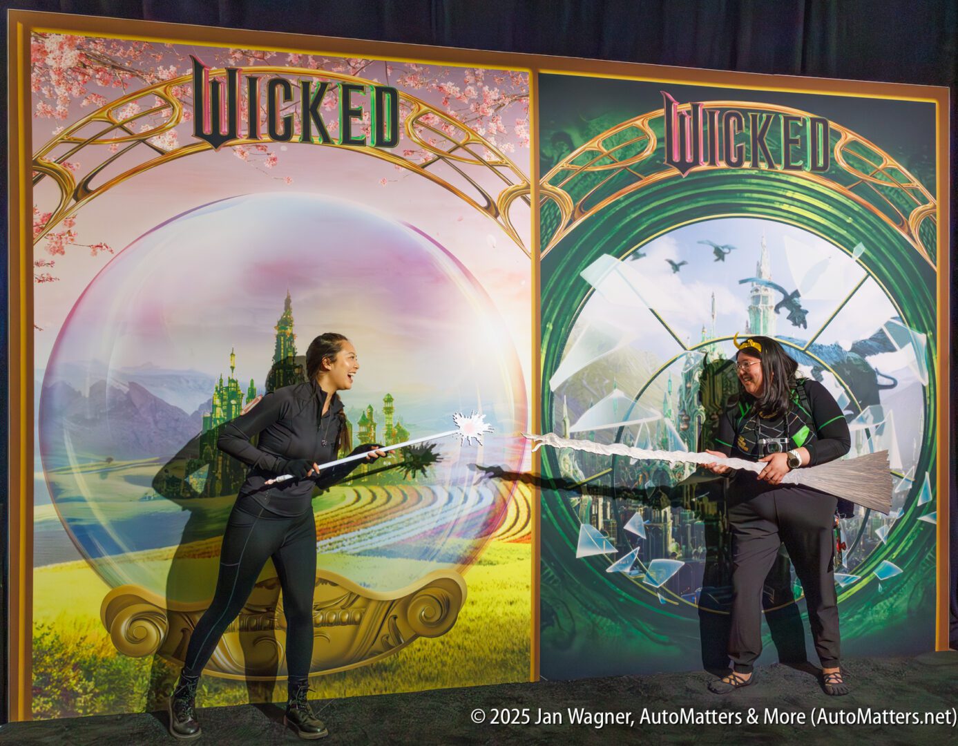 Two people pose in front of large "Wicked" movie posters, playfully acting as if they are dueling with broomsticks and a prop lightning bolt.