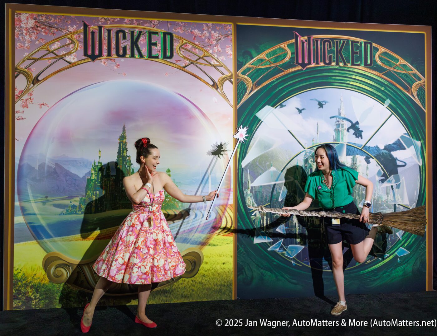 Two women pose in front of "Wicked" themed backdrops; one is dressed in a pink dress holding a wand, the other in green holding a broom as if riding it.