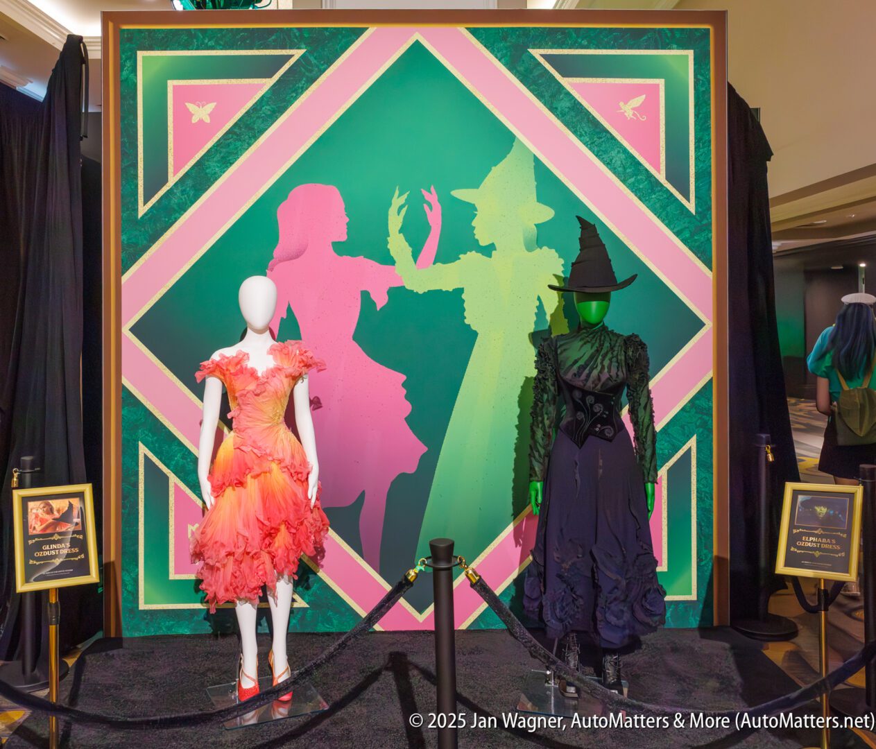 Two mannequins display costumes in front of a colorful backdrop featuring silhouettes of two women, one in a witch hat. Framed placards are positioned on either side of the display.