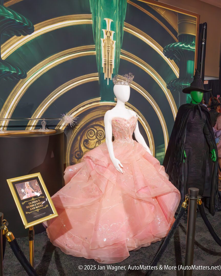 A mannequin wearing a pink, sparkly ball gown and crown stands on display next to a mannequin in a dark cloak, with a decorative backdrop and an information sign.
