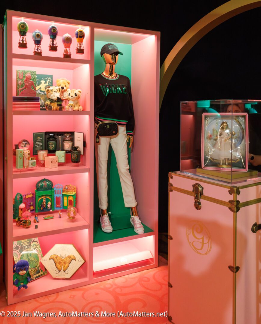 A display features shelves with various toys, candles, and collectibles, a mannequin in casual attire, and a glass case with a decorative item, all set against pink and gold accents.