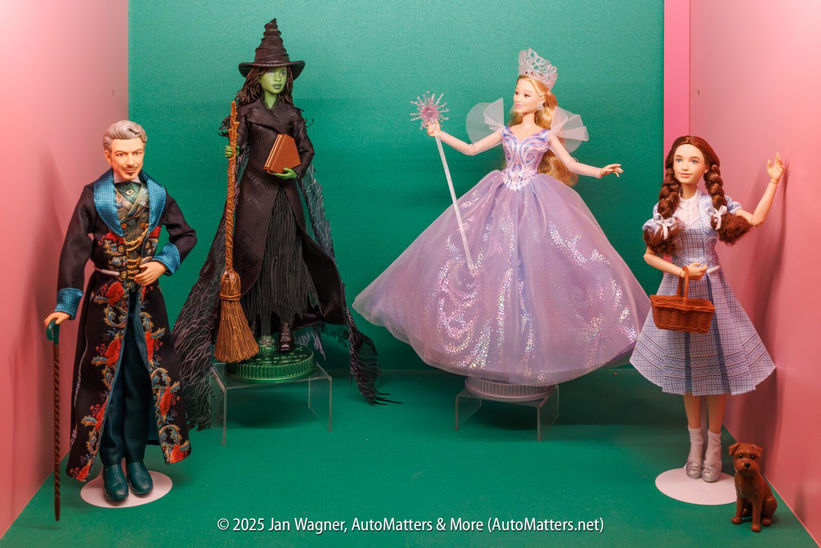 Four dolls representing characters from "The Wizard of Oz"—the Wizard, Wicked Witch, Glinda, and Dorothy with Toto—stand in front of a green and pink background.