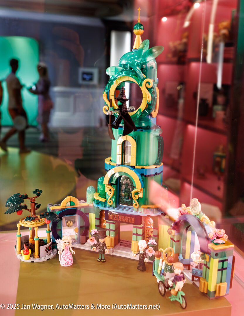 A LEGO display featuring a colorful, whimsical building with minifigures and small accessories, showcased in a glass case with reflections and background exhibits visible.