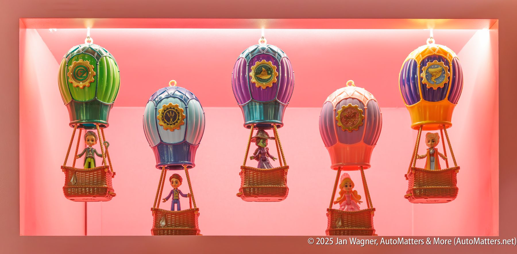 Five toy hot air balloons with dolls inside are displayed in a lit rectangular case with a pink background.