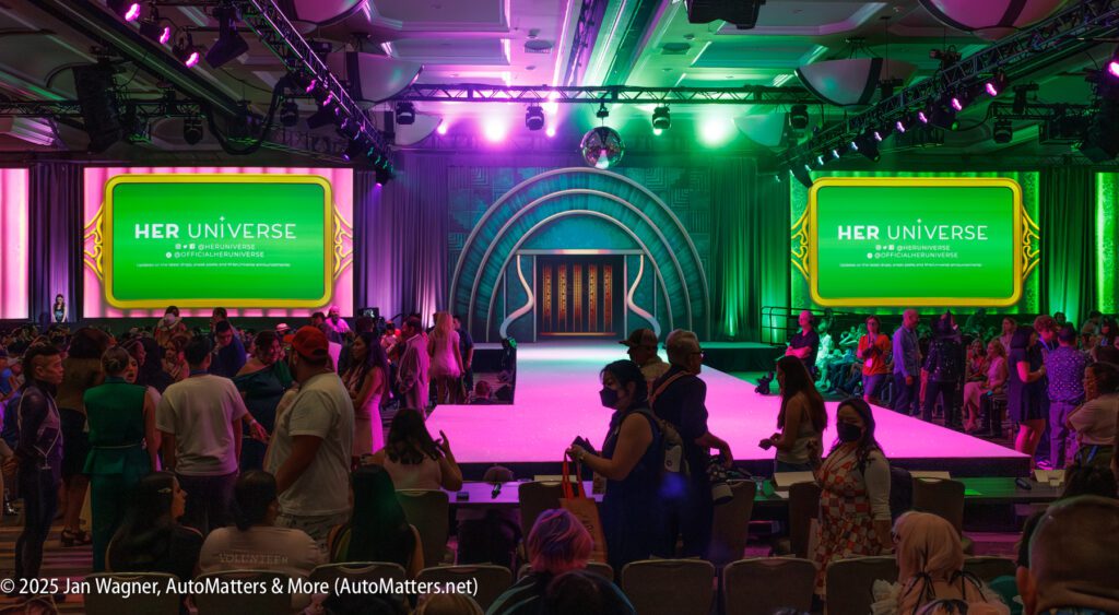 A large crowd gathers in front of a brightly lit stage with green "Her Universe" signage at a convention or event inside a spacious hall.