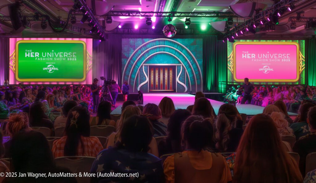Audience seated in a ballroom watching the Her Universe Fashion Show 2023, with two large screens displaying event graphics on either side of a lit stage.