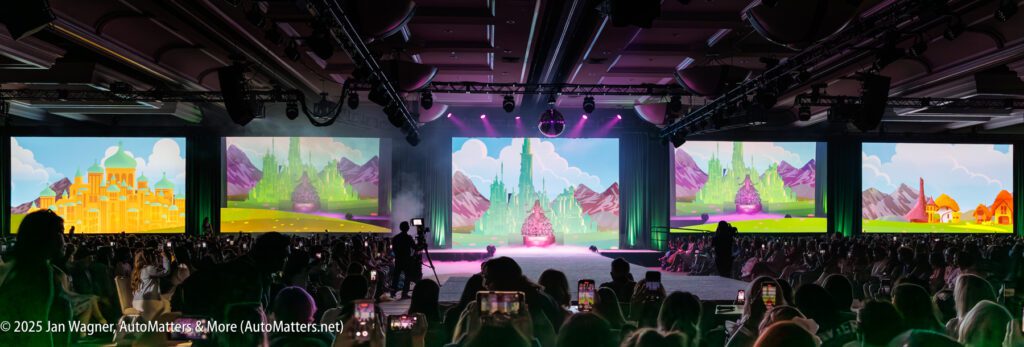 A large audience faces a stage with multiple wide screens displaying colorful animated castles and landscapes in a convention hall setting.