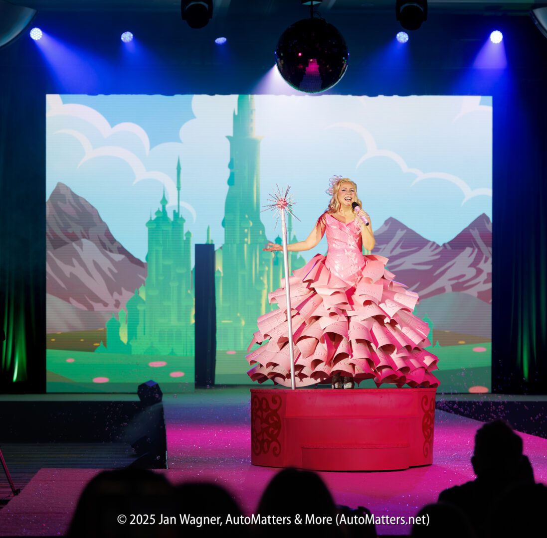 A performer in a pink dress and crown stands on a red platform, holding a wand, with a castle scene displayed on a screen behind her.