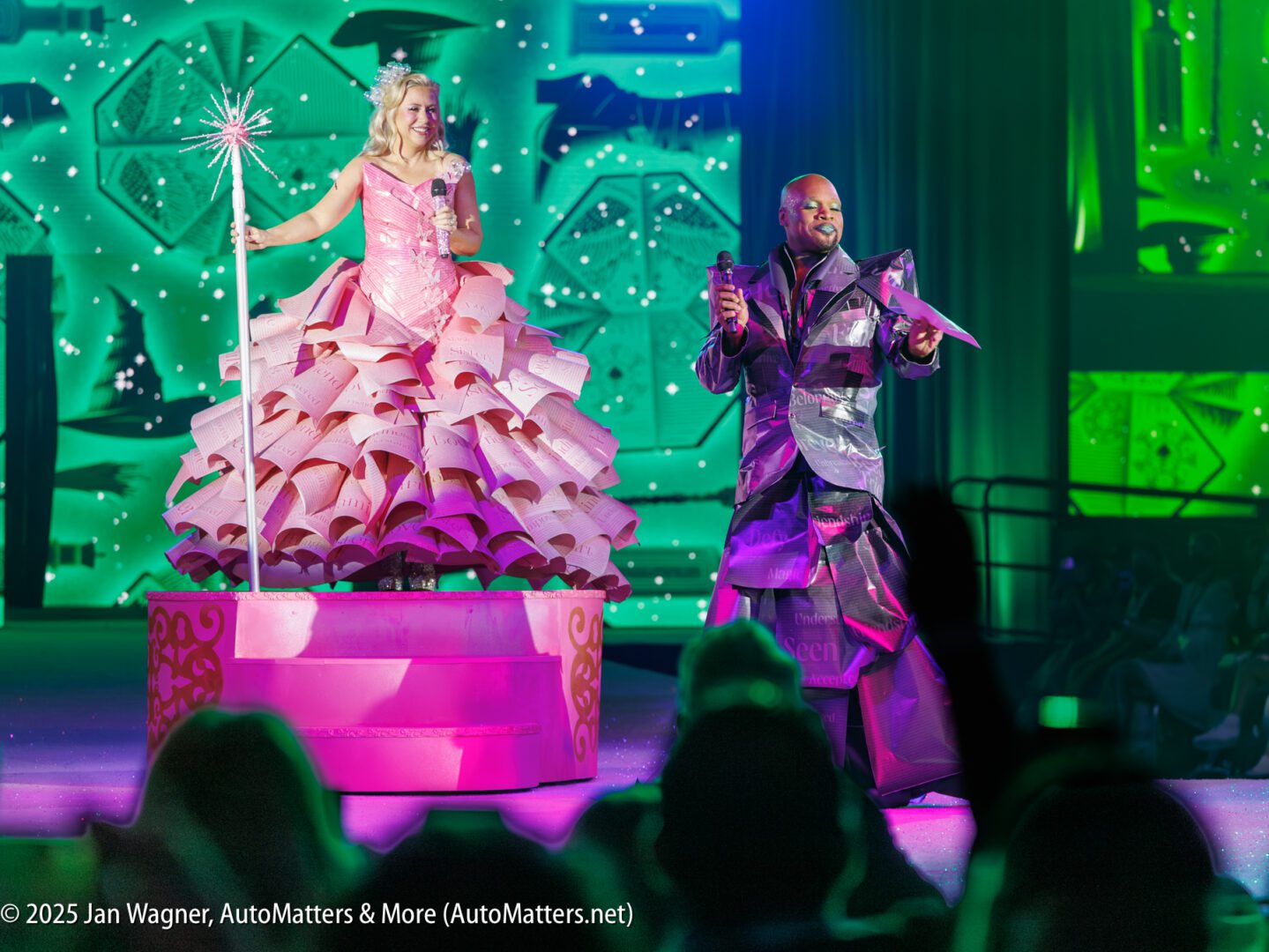 A woman in a pink, elaborate dress stands on a float holding a wand, next to a man in a metallic costume singing on stage.