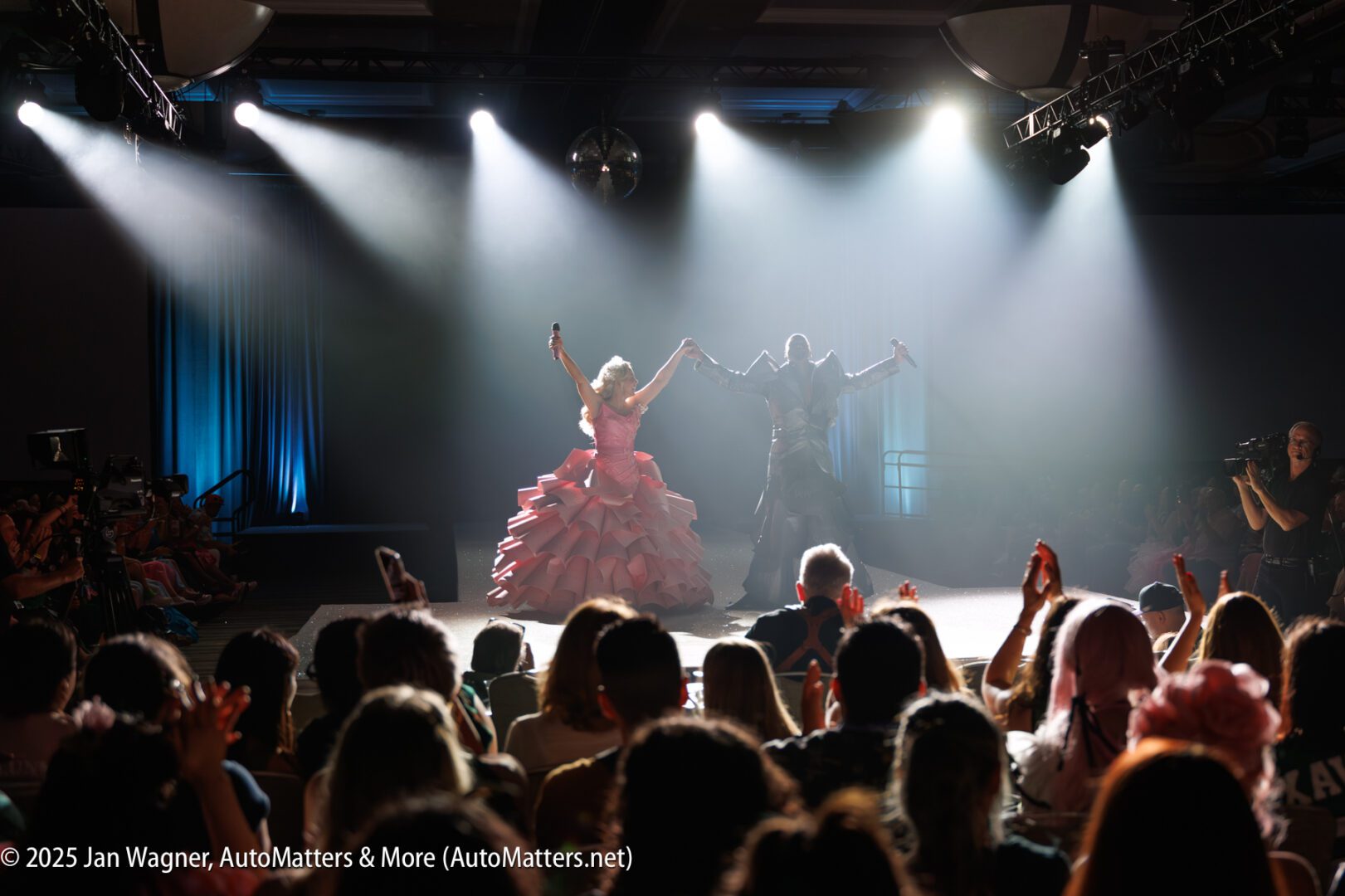 Two performers, one in a pink dress and one in a dark outfit, stand on stage holding hands as the audience applauds under bright stage lights.