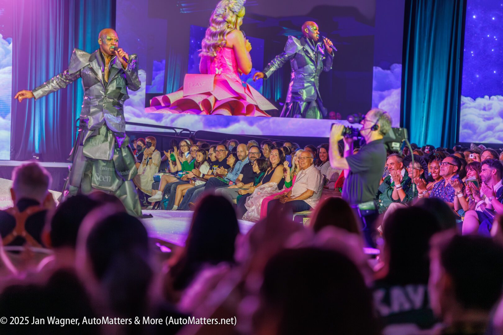A performer sings on stage in front of an audience, with a camera operator filming and a woman sitting on stage holding a clipboard. The event appears lively and well-attended.