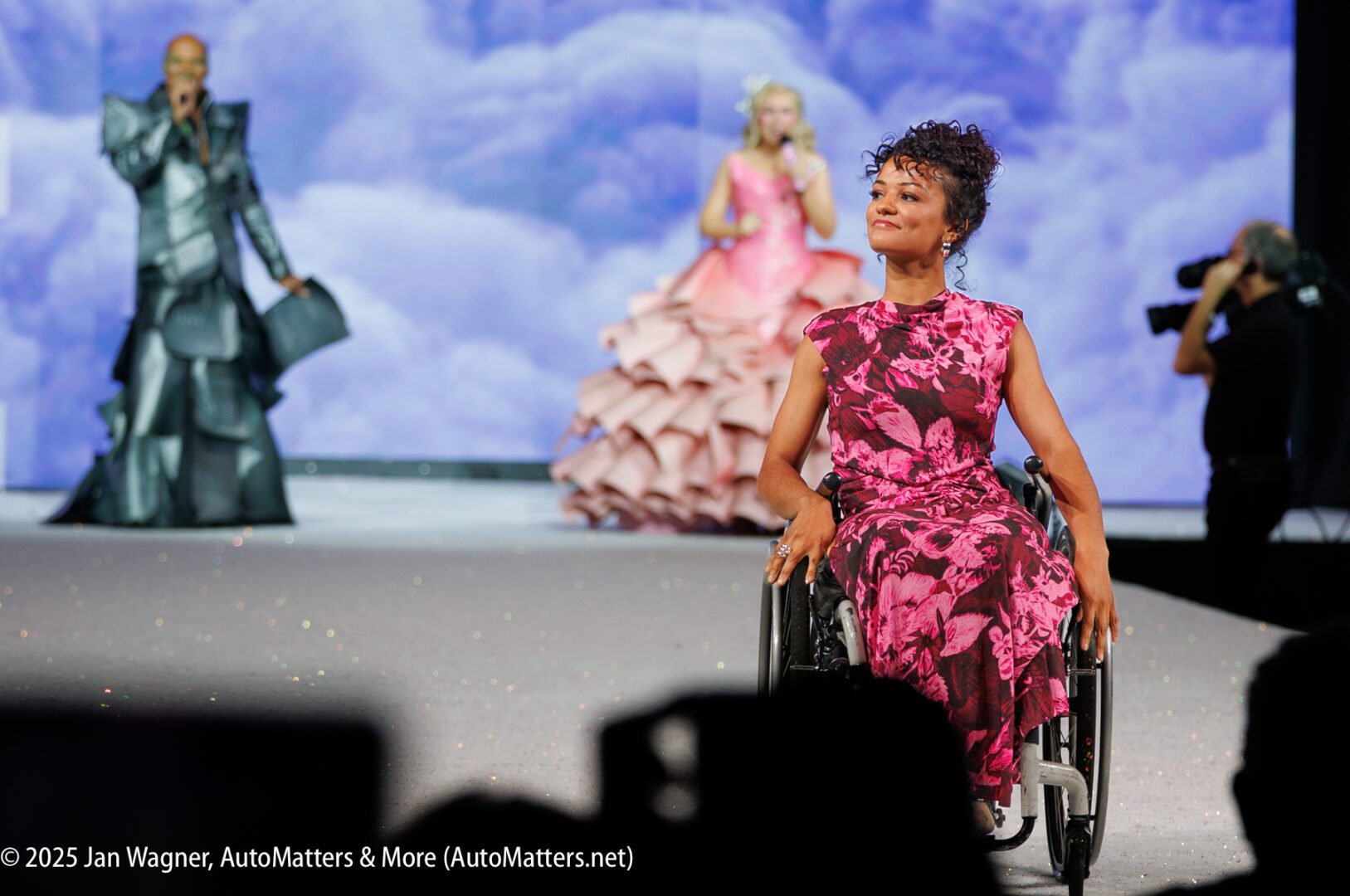 A woman in a pink floral dress uses a wheelchair on a runway, with two standing models and a photographer in the background.