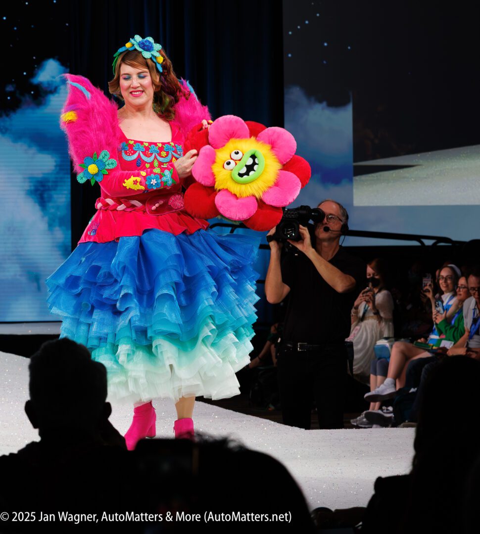 A woman in a colorful, layered dress with pink wings walks down a runway holding a plush flower toy, while a photographer captures the moment and the audience watches.