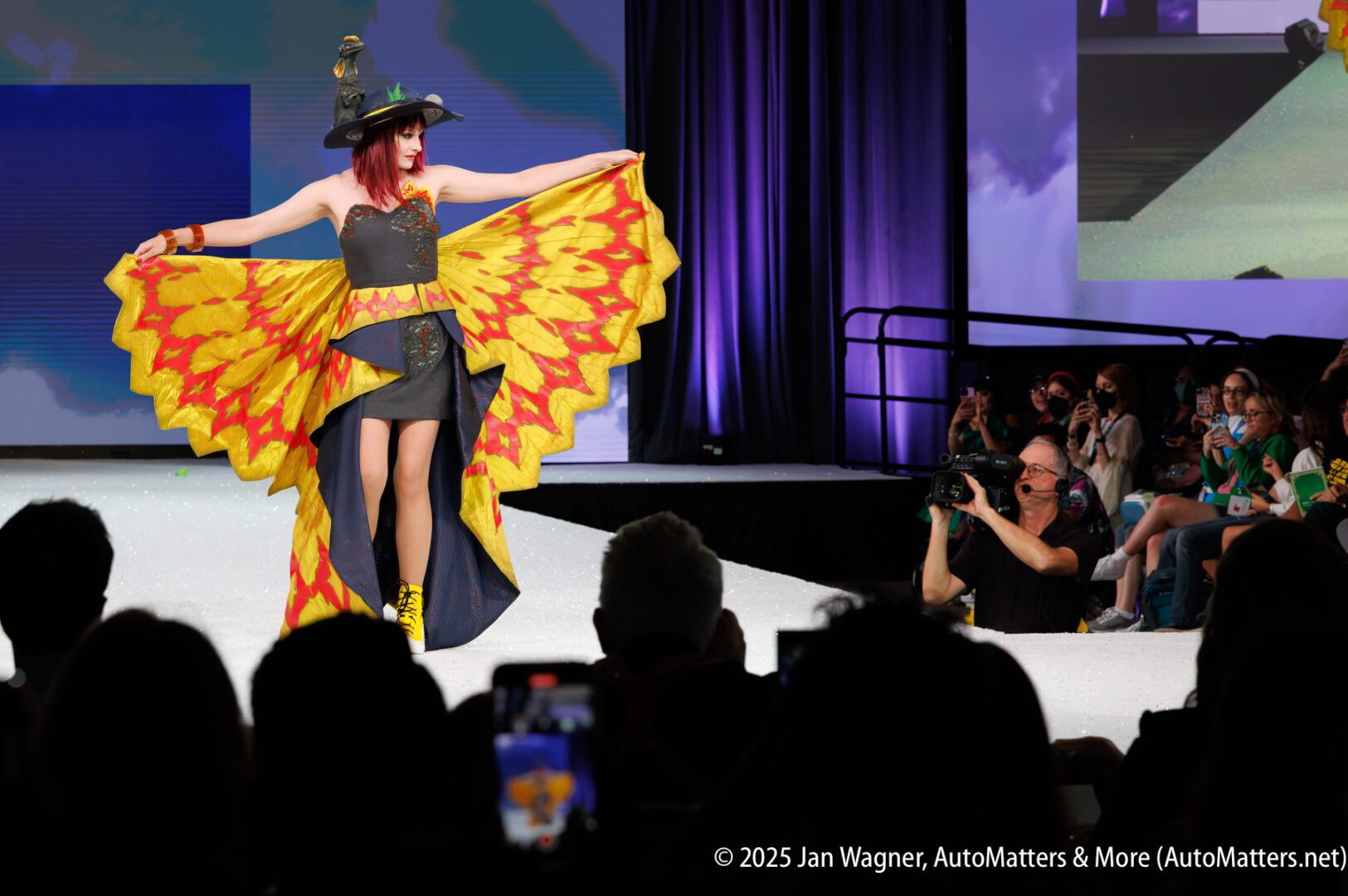 A woman in a black and yellow dress with wing-like sleeves poses on a runway, surrounded by photographers and an audience.