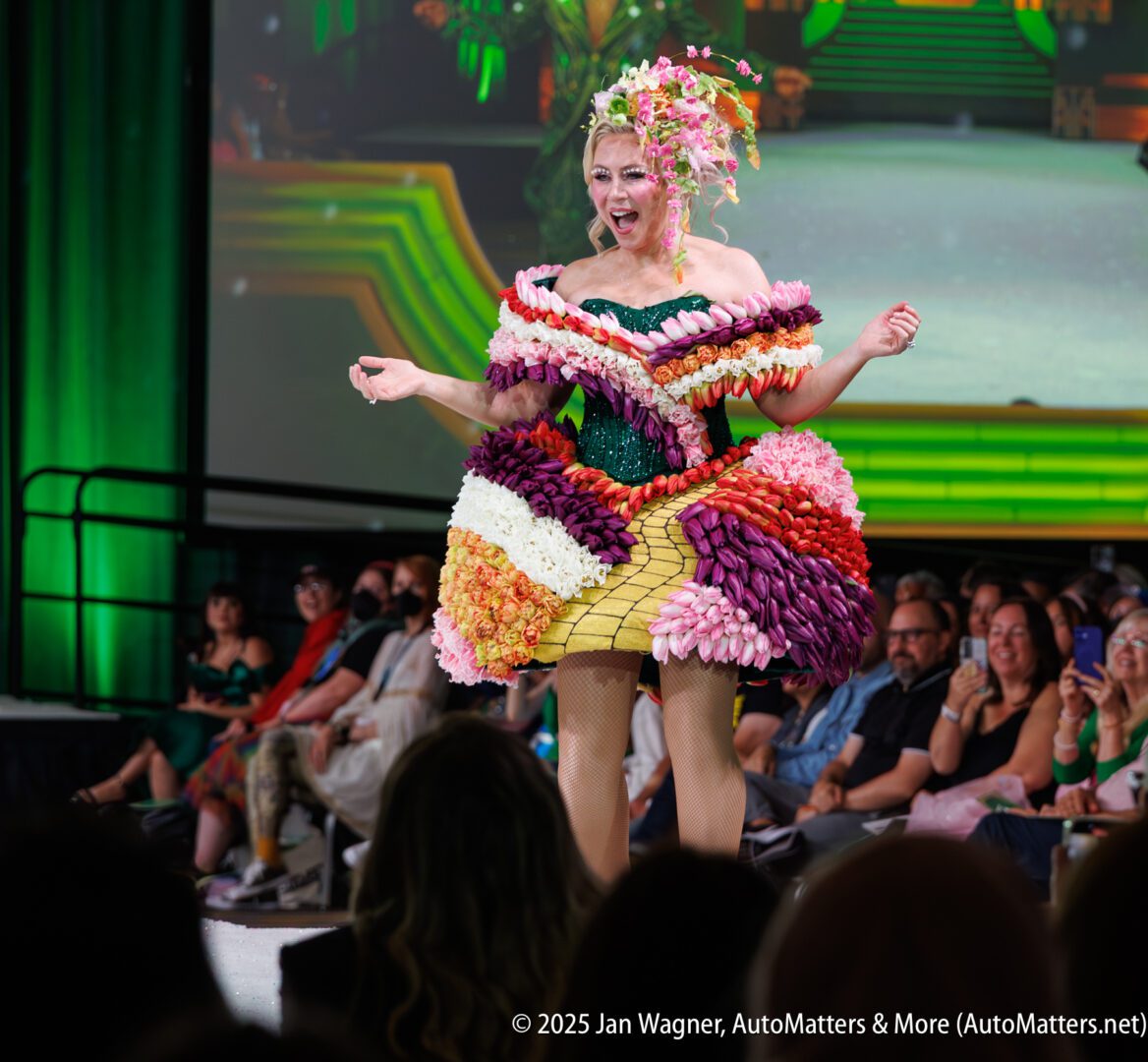 Model walks on a runway wearing a colorful, elaborate floral-themed dress and headpiece, with audience watching in the background.
