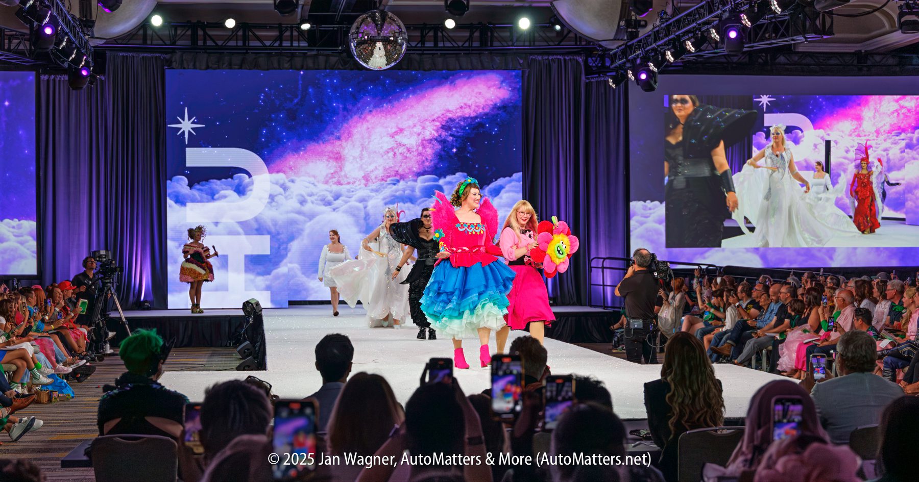 A fashion show runway features diverse models in colorful and white outfits, walking in front of a cosmic-themed backdrop, with an audience seated on both sides.