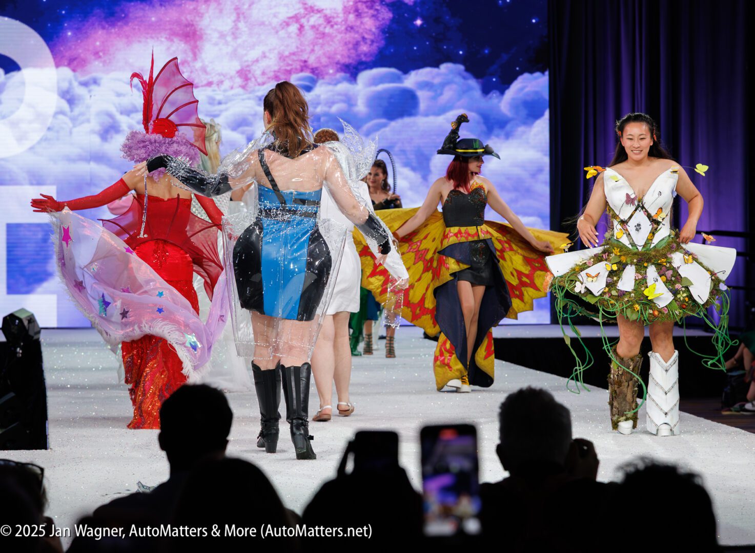 A group of women wearing elaborate, colorful costumes walk on a fashion runway, with some facing forward and others turned away. Audience members are visible in the foreground.