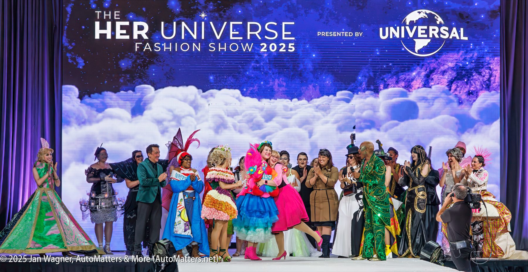 A group of people in colorful, elaborate costumes stand on stage at the Her Universe Fashion Show 2025, with a cloud-themed backdrop and audience applauding.