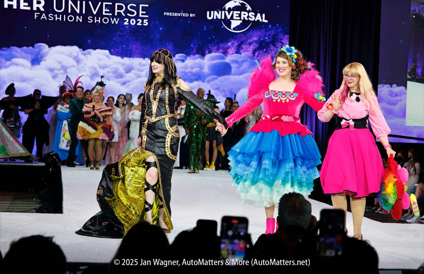 Three women in elaborate, colorful costumes walk hand-in-hand on a fashion show runway, with other costumed participants and an audience visible in the background.
