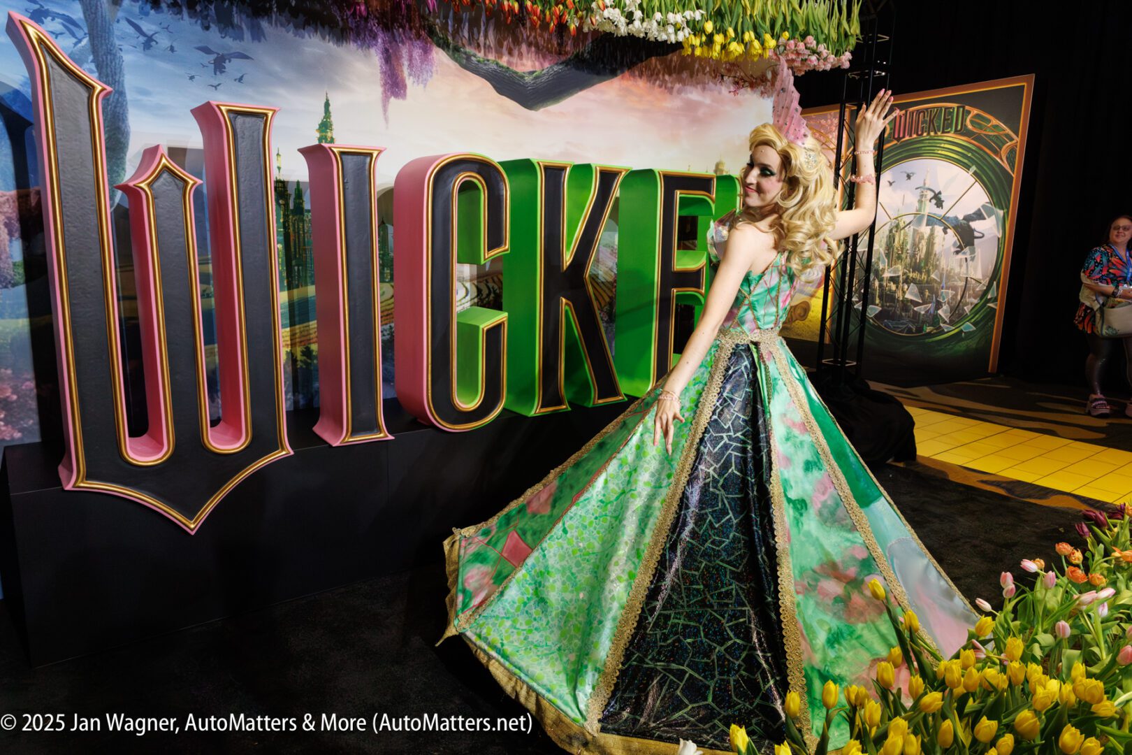 A person in a green and gold gown poses in front of a large "WICKED" sign at an event, surrounded by flowers and themed decorations.