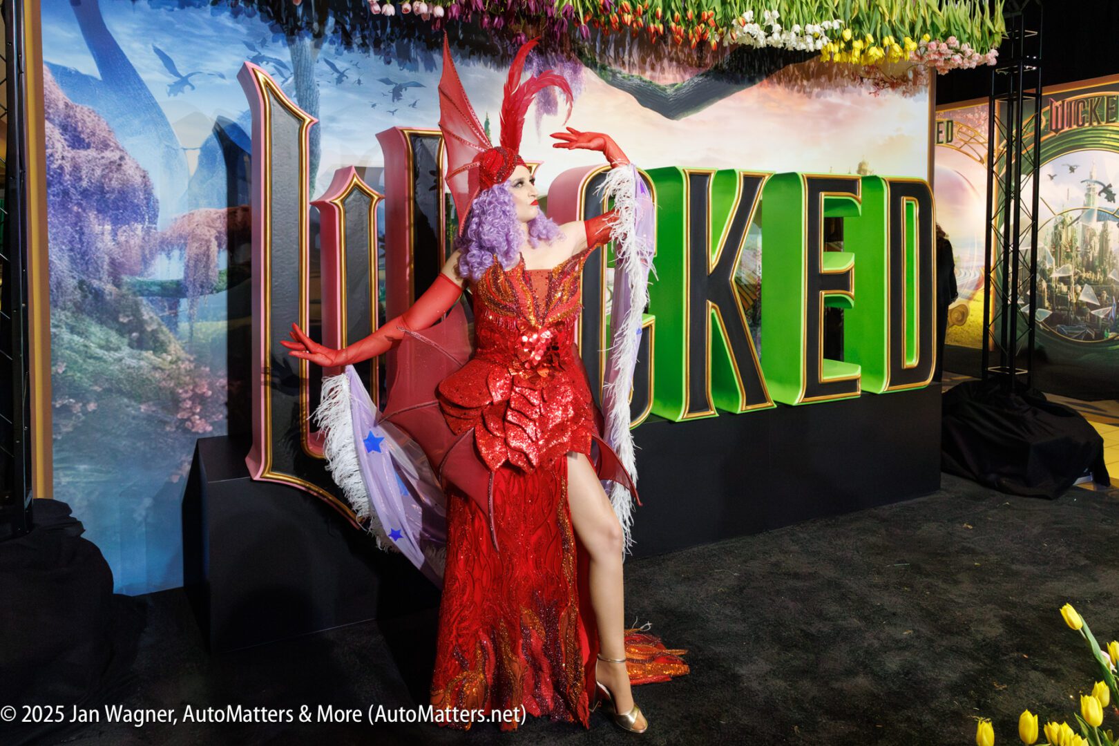 A person in a red and purple feathered costume poses in front of a large "WICKED" sign at a themed event or premiere.