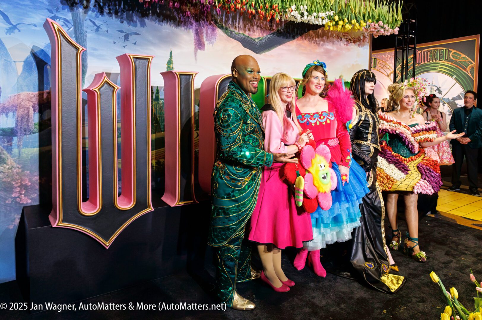 A group of people in colorful costumes pose for a photo in front of a large "Wicked" sign at an event.