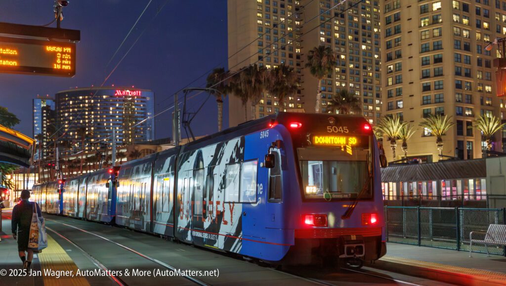 A blue and red trolley labeled "Downtown SD" stops at a lit outdoor station in an urban area with tall buildings and palm trees at dusk.
