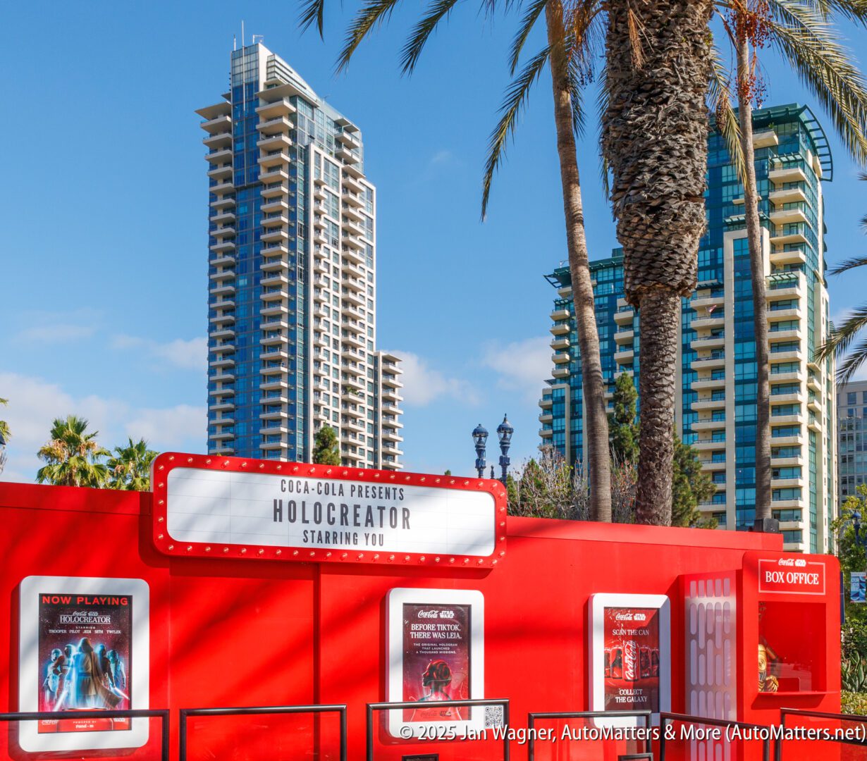 A red box office booth with a marquee sign reads "Holocreator Starring You," set against tall modern buildings and palm trees on a sunny day.