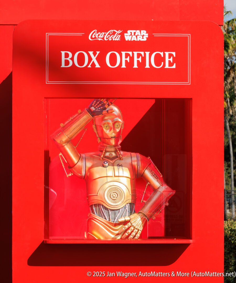 A gold humanoid robot is posed behind glass in a red box office booth with Coca-Cola and Star Wars logos above.