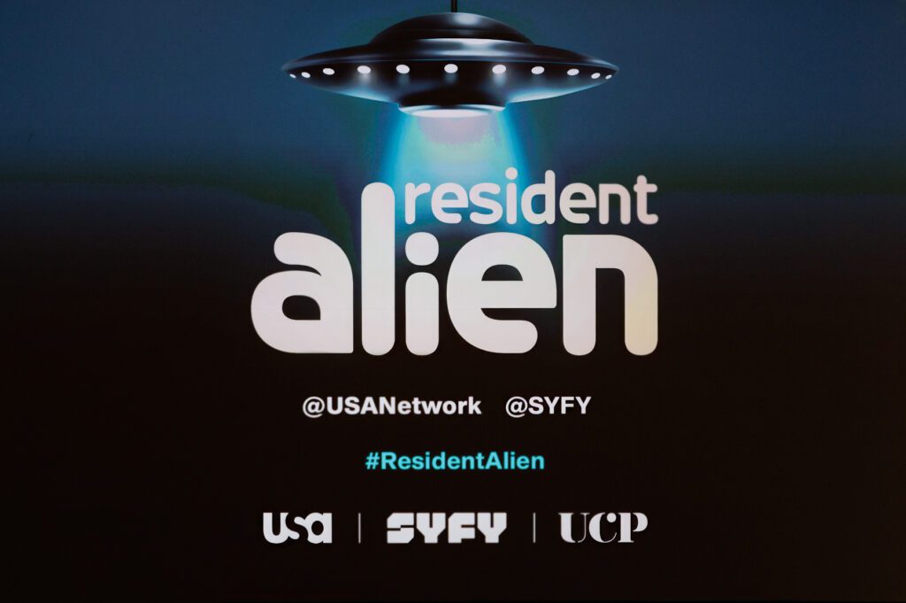 A graphic for "Resident Alien" shows a UFO beaming light over the title, with social media handles and logos for USA, SYFY, and UCP at the bottom.