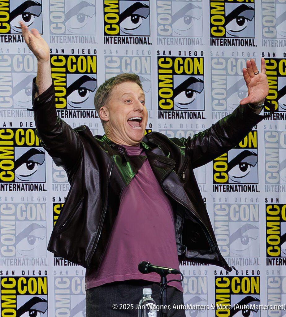 A man in a leather jacket enthusiastically raises his arms on stage at San Diego Comic-Con International in front of a branded backdrop.