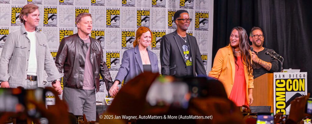 A group of six people stand together on stage at San Diego Comic-Con, with Comic-Con logos on the backdrop and an audience taking photos.