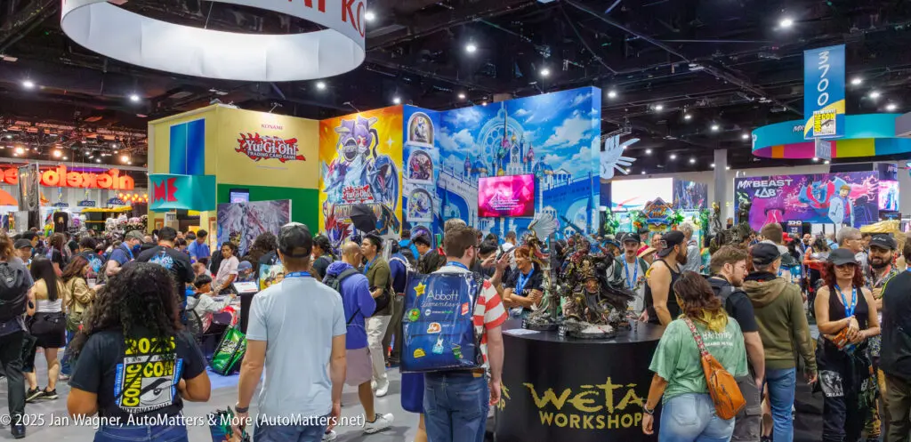 A crowded convention hall features colorful booths, including Weta Workshop and Anime Expo, with attendees walking, browsing, and taking photos.