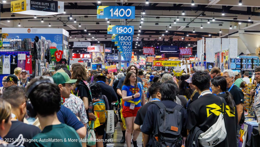 A large crowd walks through crowded aisles at a convention with colorful signs, vendor booths, and banners for brands like Marvel visible in the background.