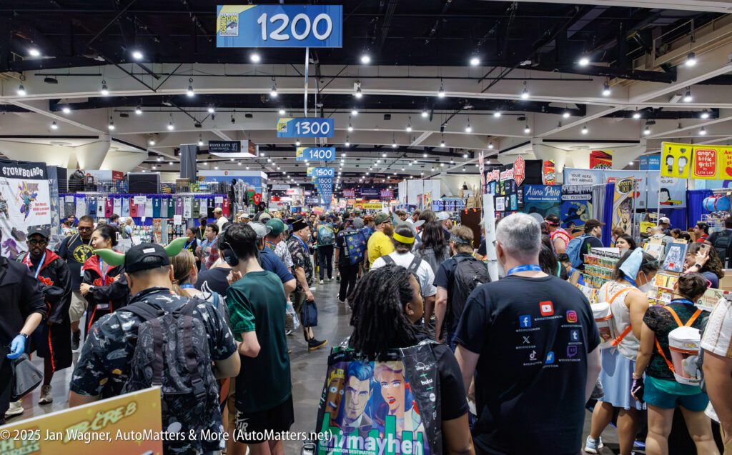 A crowded convention hall with attendees walking among vendor booths under blue aisle signs labeled 1200 and 1300.