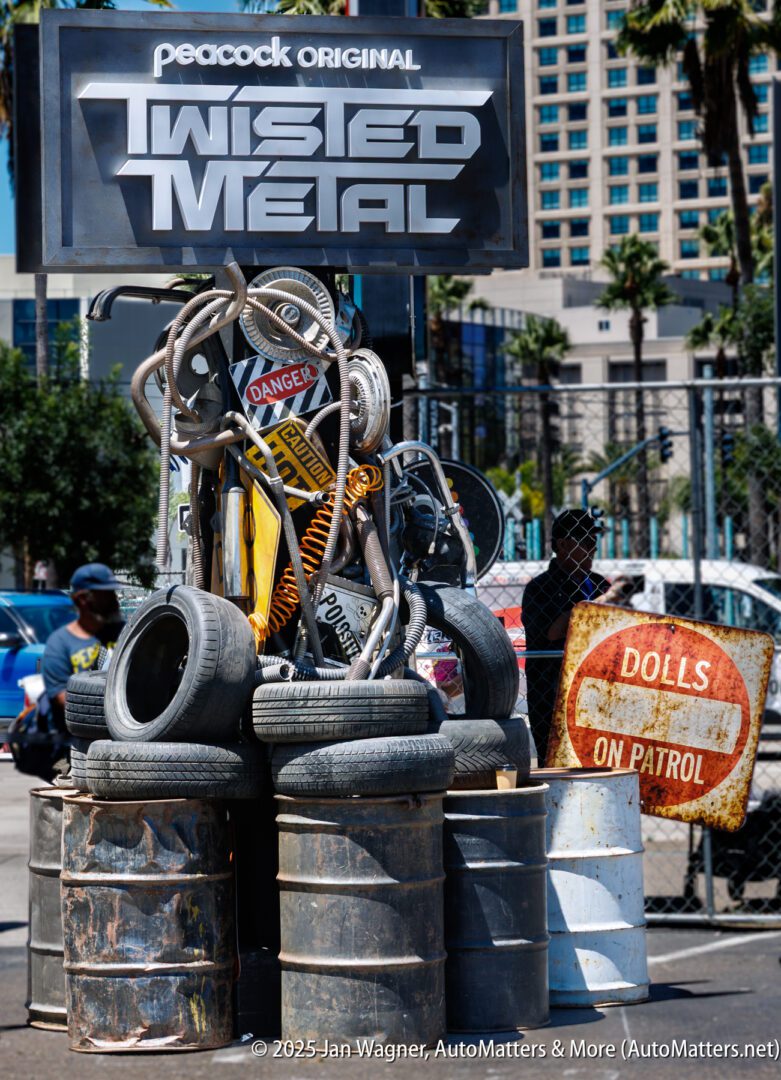 A promotional display for "Twisted Metal" featuring stacked tires, metal scraps, warning signs, and a "Dolls on Patrol" sign, set outdoors with buildings in the background.