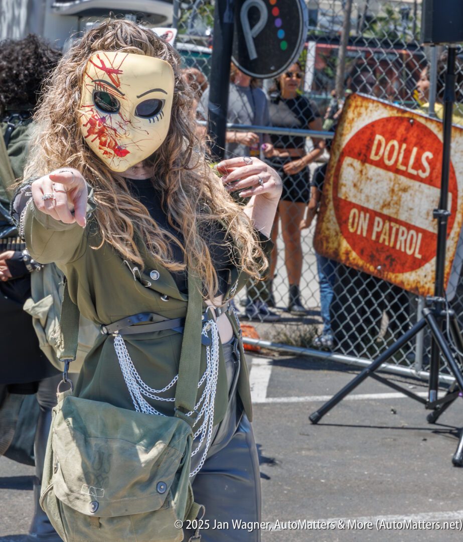 Person in olive green outfit and a half-damaged doll mask points toward the camera at an outdoor event; a "DOLLS ON PATROL" sign is visible in the background.