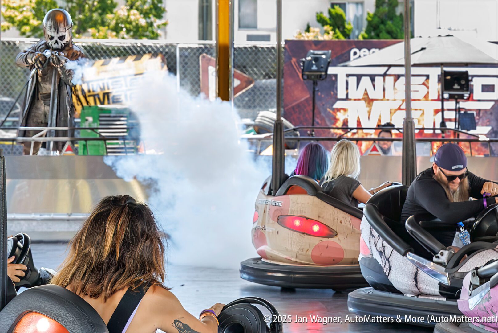Several people ride bumper cars while a performer in costume releases smoke on a platform nearby at an amusement event.